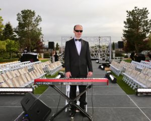 Santon is standing behind his keyboard on an outdoor stage. He is in a black tuxedo and wearing black sunglasses